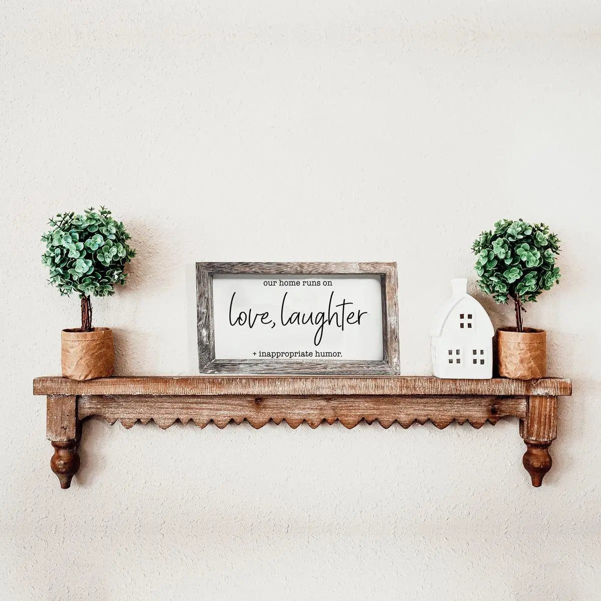 Wooden shelf with decorative items including a framed sign, potted plants, and a small house-shaped object on a plain wall.