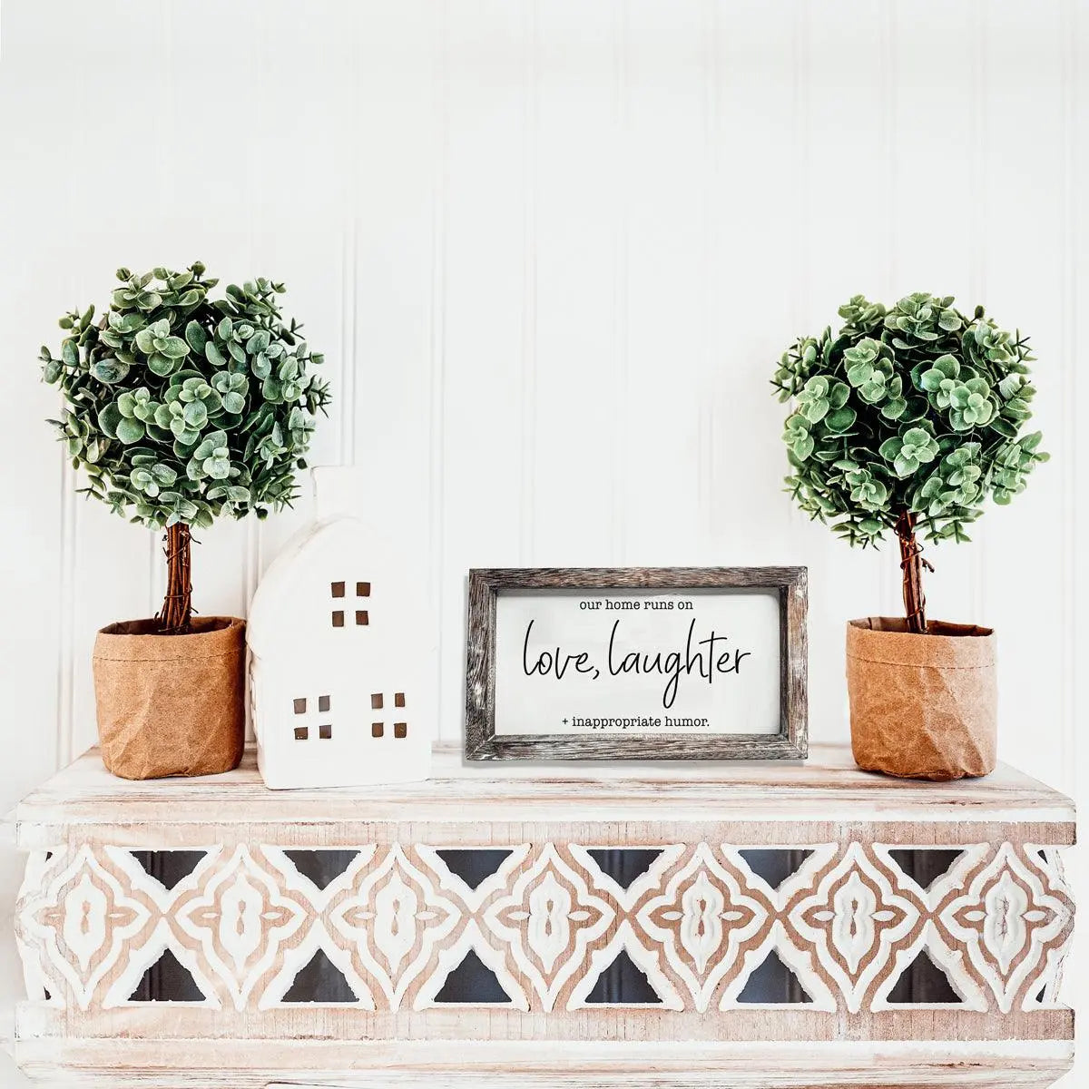 Decorative shelf with two potted plants, a small house-shaped object, and a framed quote on a white wall.