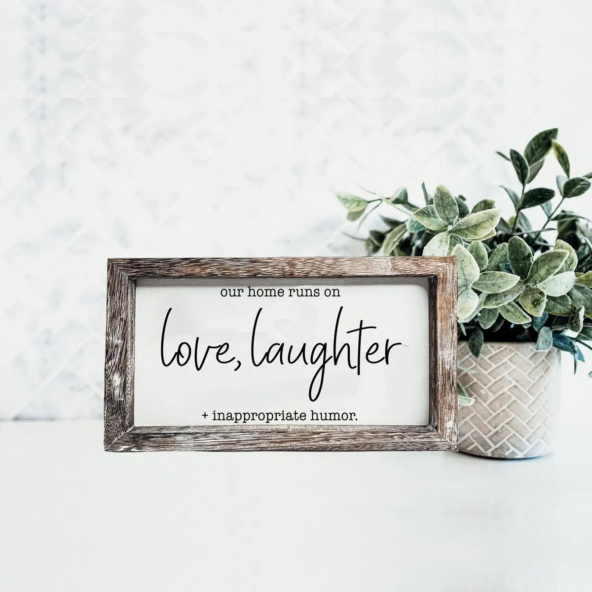 Framed sign with text 'our home runs on love, laughter + inappropriate humor' on a white wall with a plant.