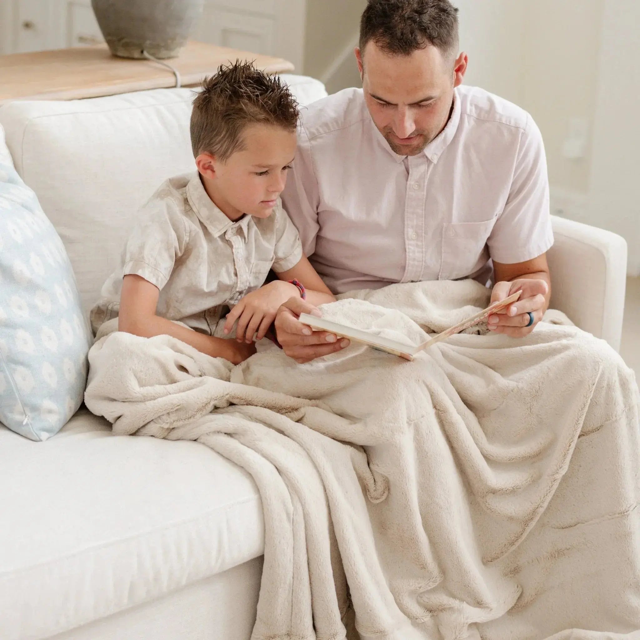 Man and child sitting on a couch under a blanket, looking at something together.