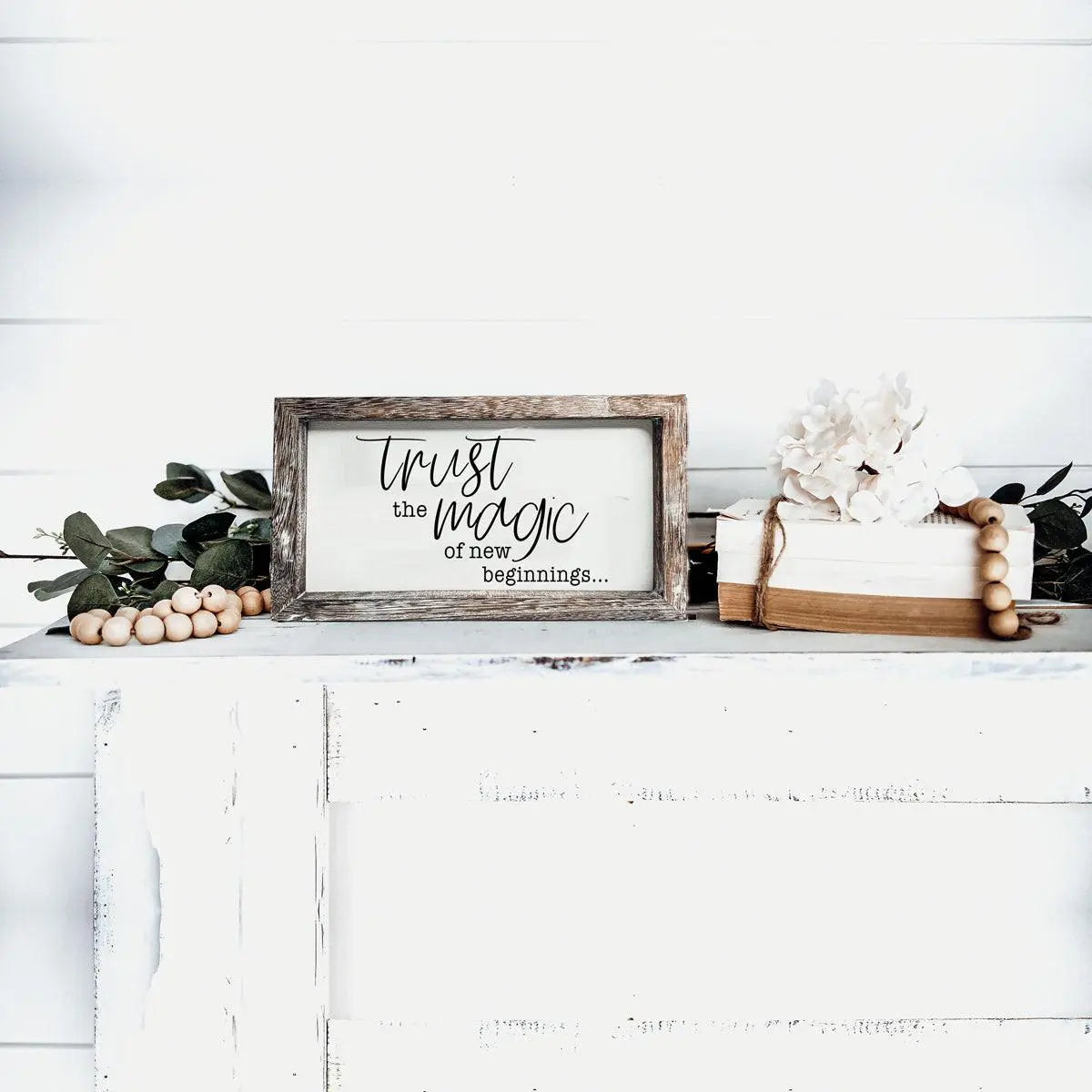 Framed sign with 'Trust the magic of new beginnings' on a white surface with decorative elements.