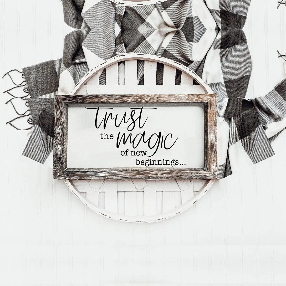 Decorative sign with 'Trust the magic of new beginnings' text on a white background with plaid fabric.