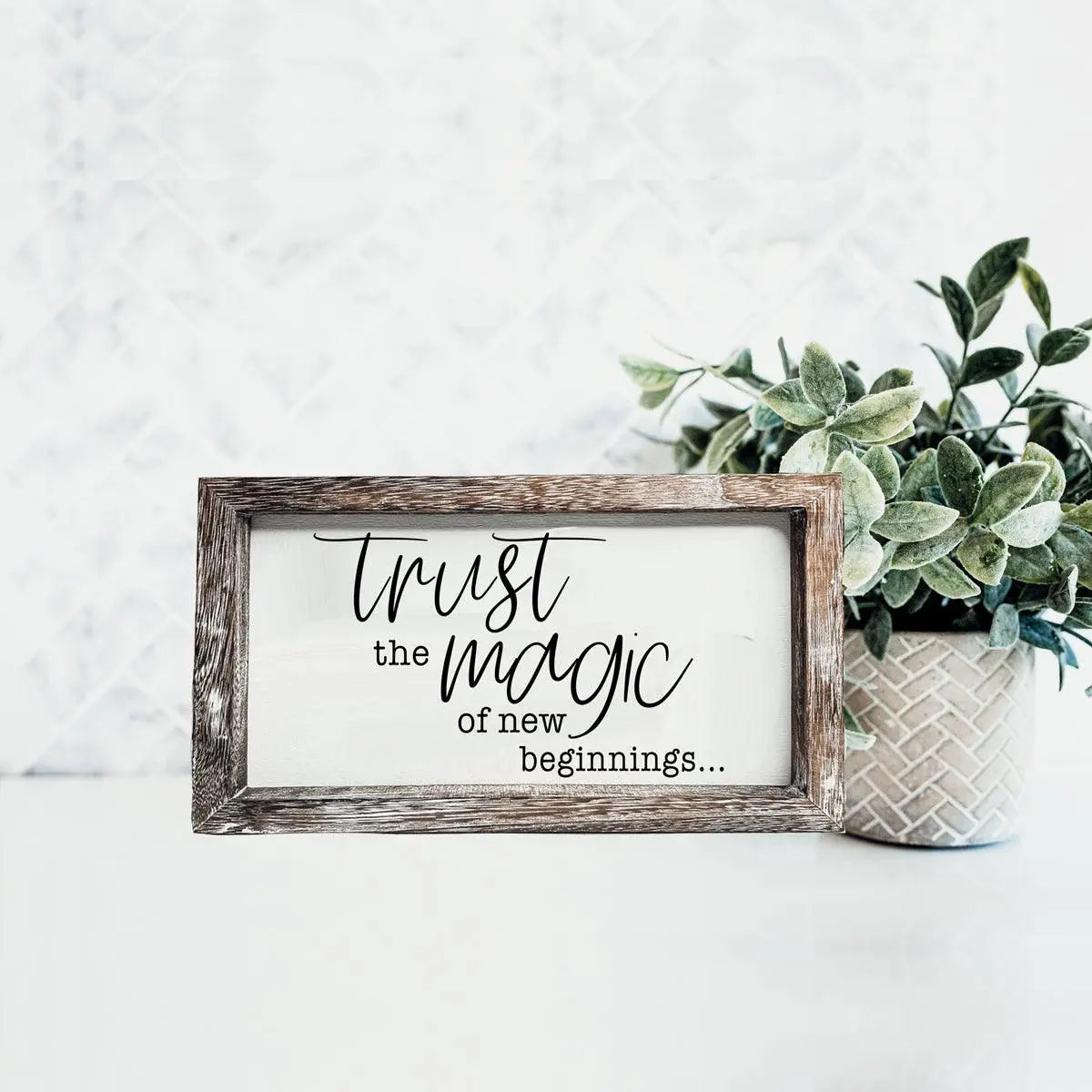 Framed sign with 'Trust the magic of new beginnings' text on a white surface with a plant.