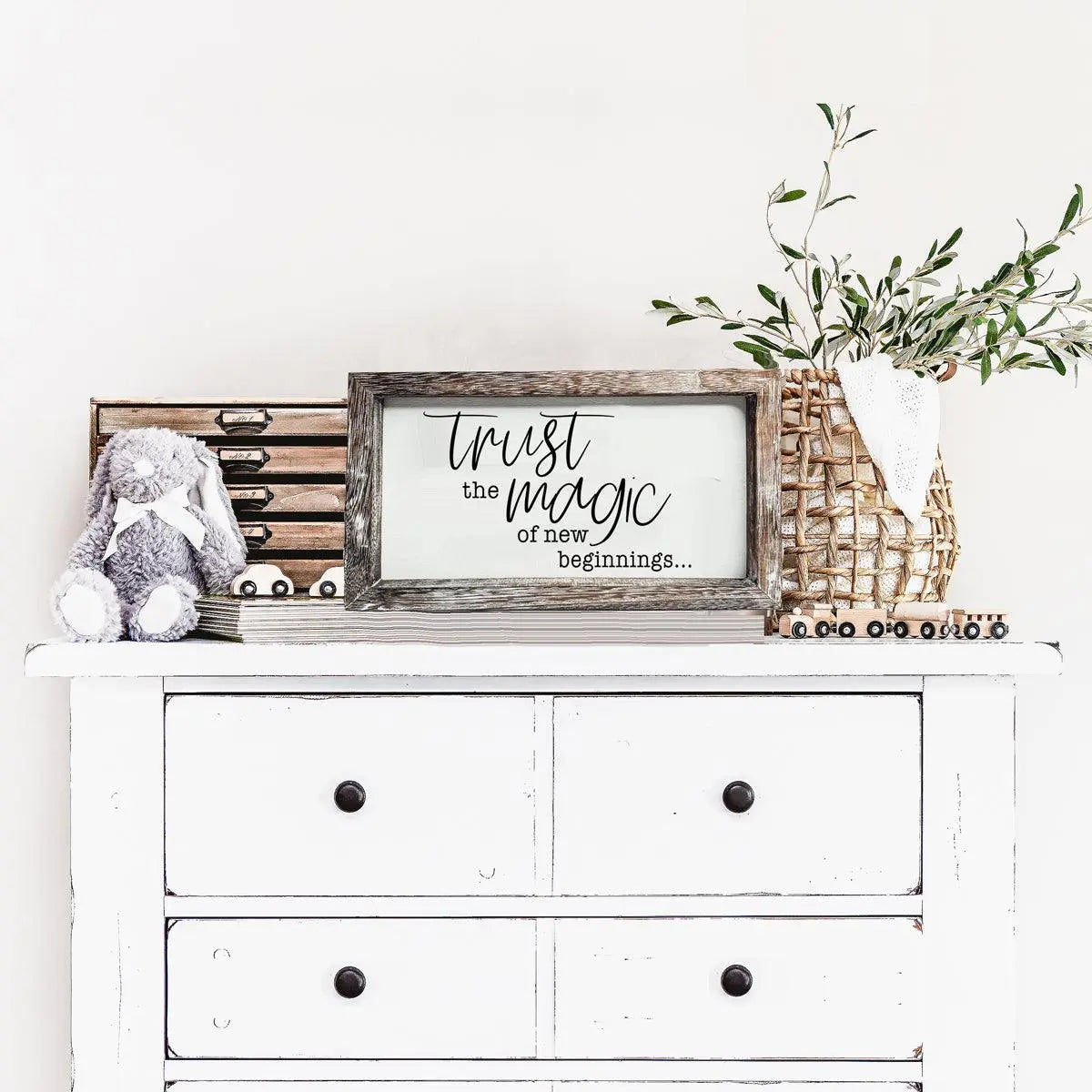 White dresser with decorative items including a framed quote, a stuffed bunny, and a plant.