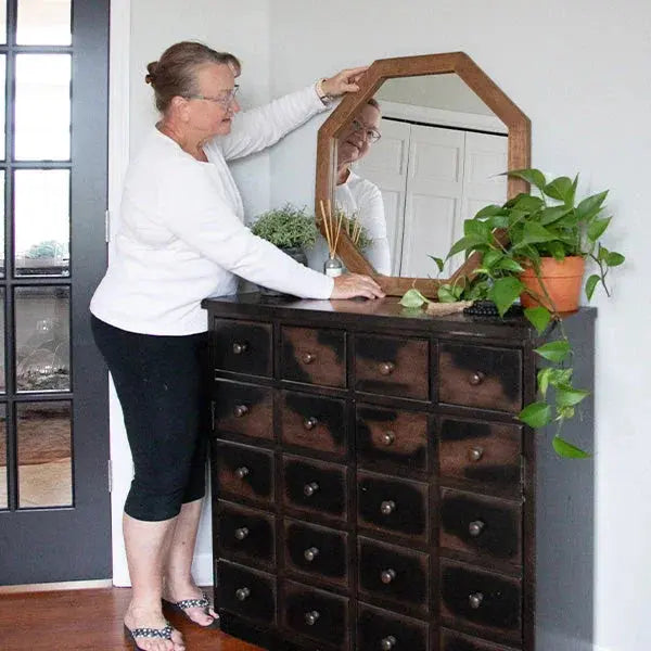 Woman arranging hexagon mirror on rustic apothecary cabinet with green plant, farmhouse decor