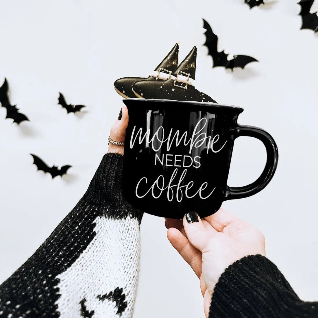 Black mug with 'Mommy Needs Coffee' text held by a person against a white background with black bats.
