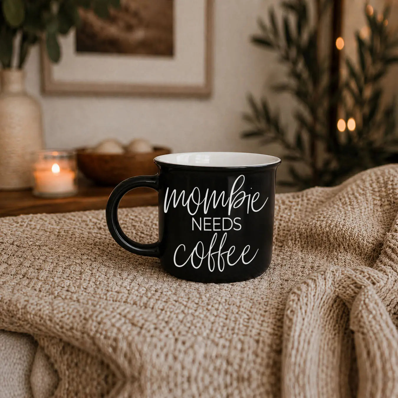 Black mug with 'mommy needs coffee' text on a textured surface with a cozy background.