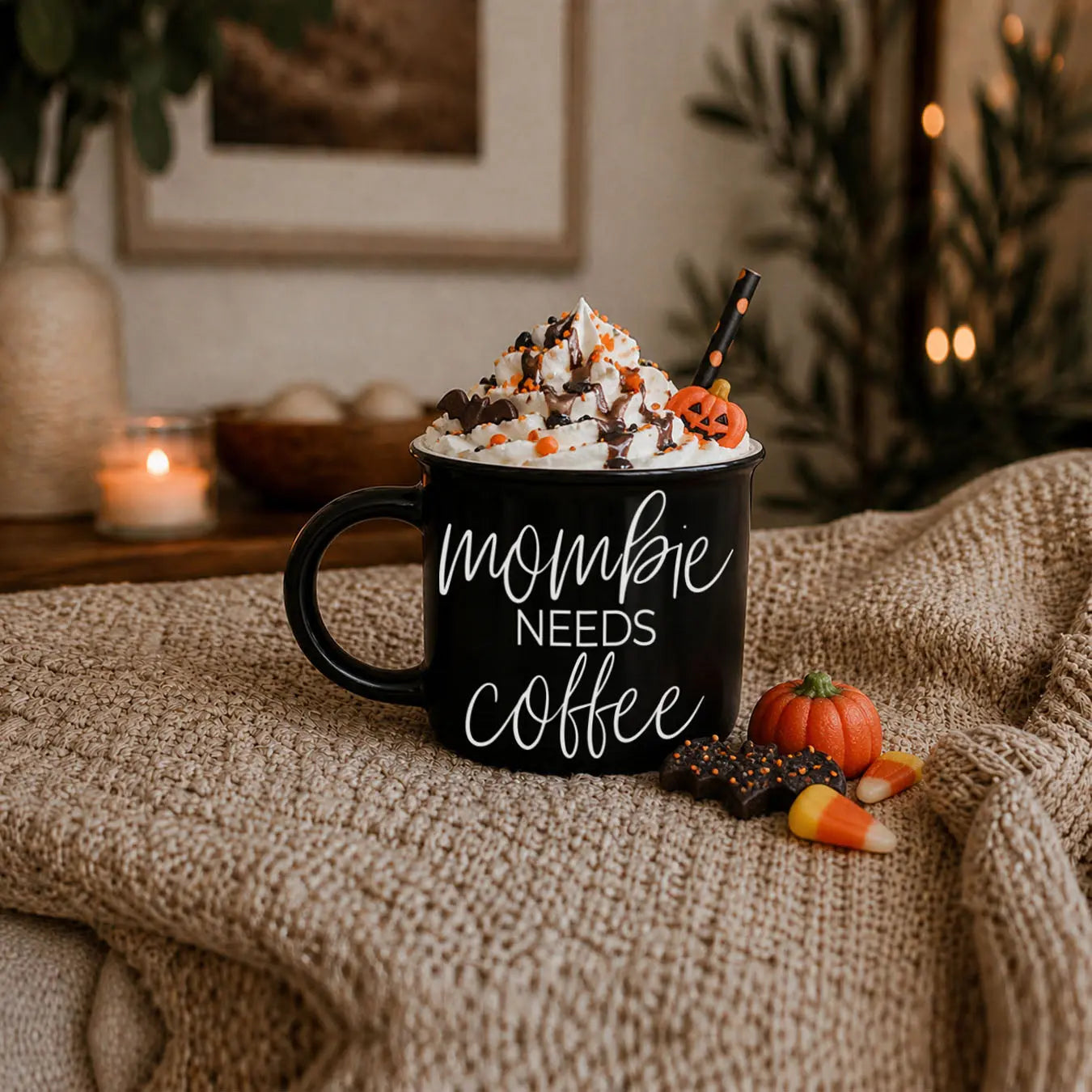 Black mug with 'mombie needs coffee' text, topped with whipped cream and Halloween decorations, on a textured surface.