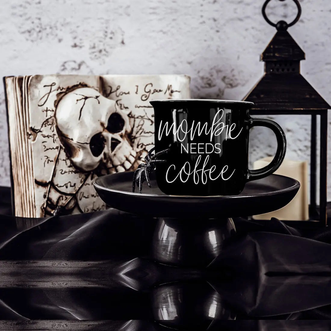 Black mug with 'mommy needs coffee' text on a saucer, skull, and candle in a dark setting