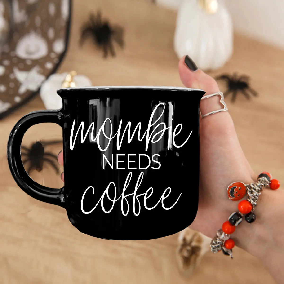Black mug with 'mommy needs coffee' text held by a hand, with Halloween decorations in the background.