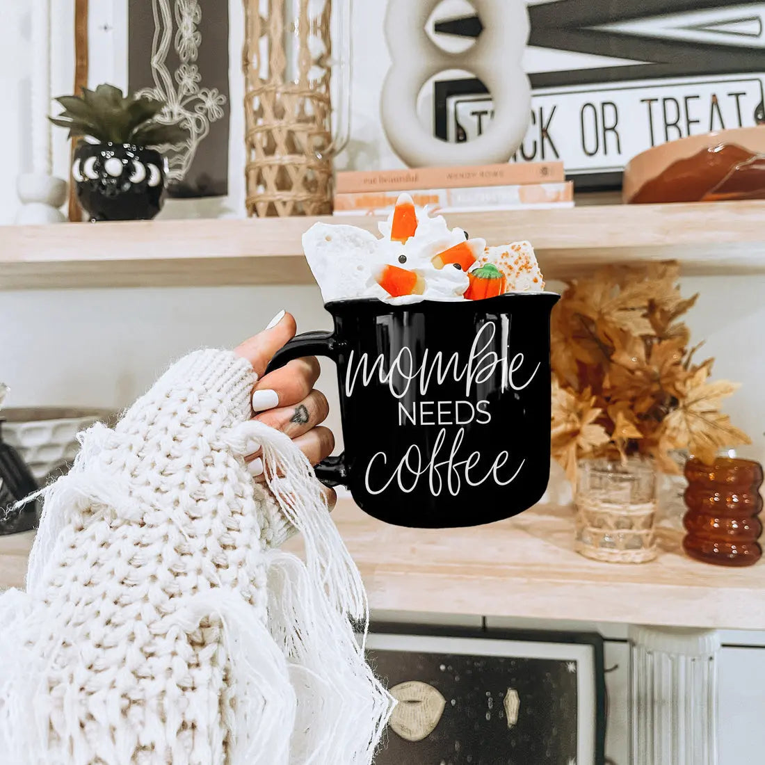 Person holding a black mug with 'mommy needs coffee' text, surrounded by decorative items on a shelf.
