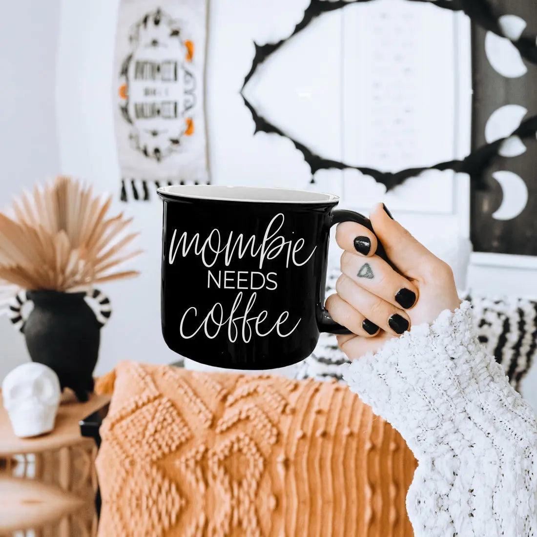 Person holding a black mug with 'mommie needs coffee' text in a cozy room.