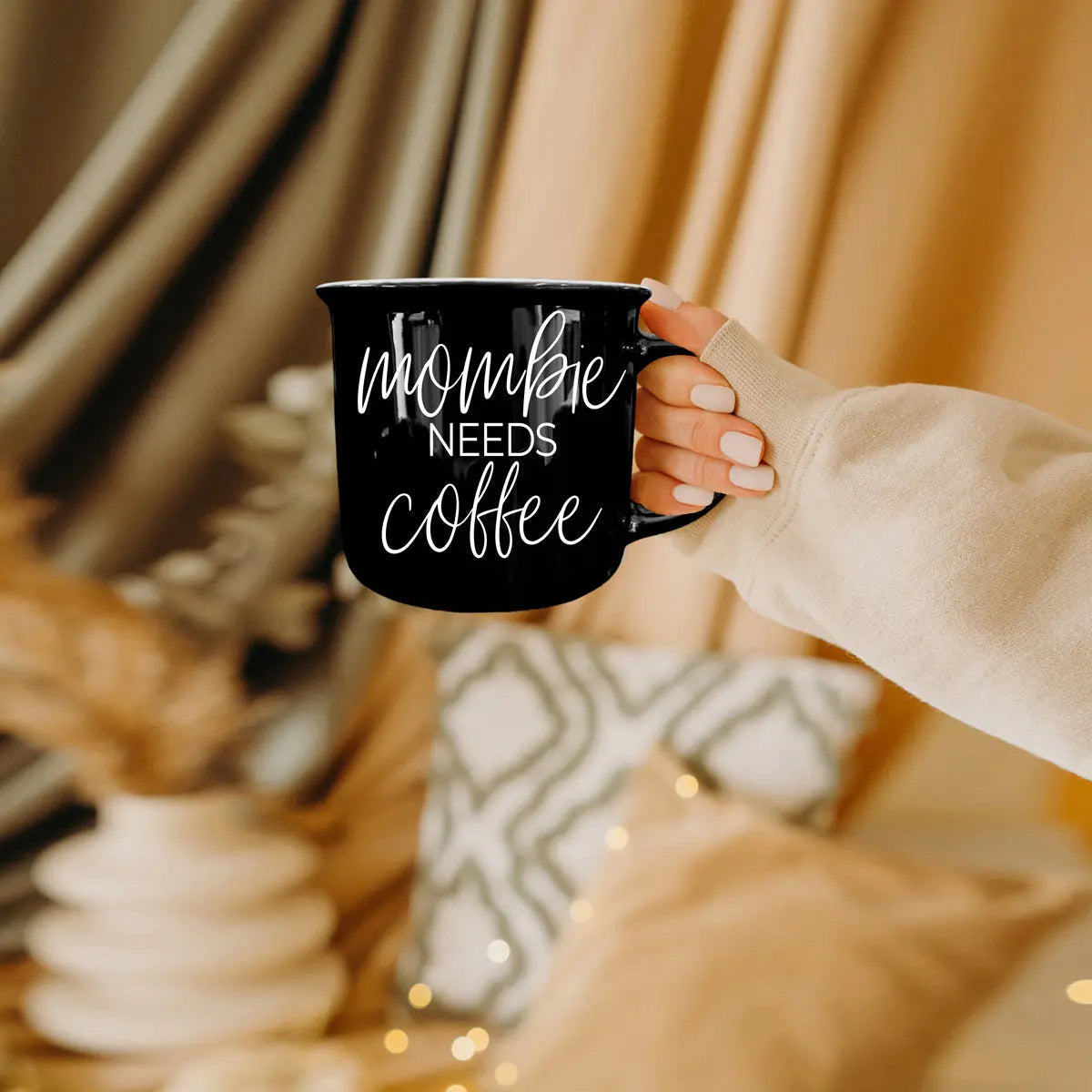 Person holding a black mug with 'mommy needs coffee' text in a cozy indoor setting.