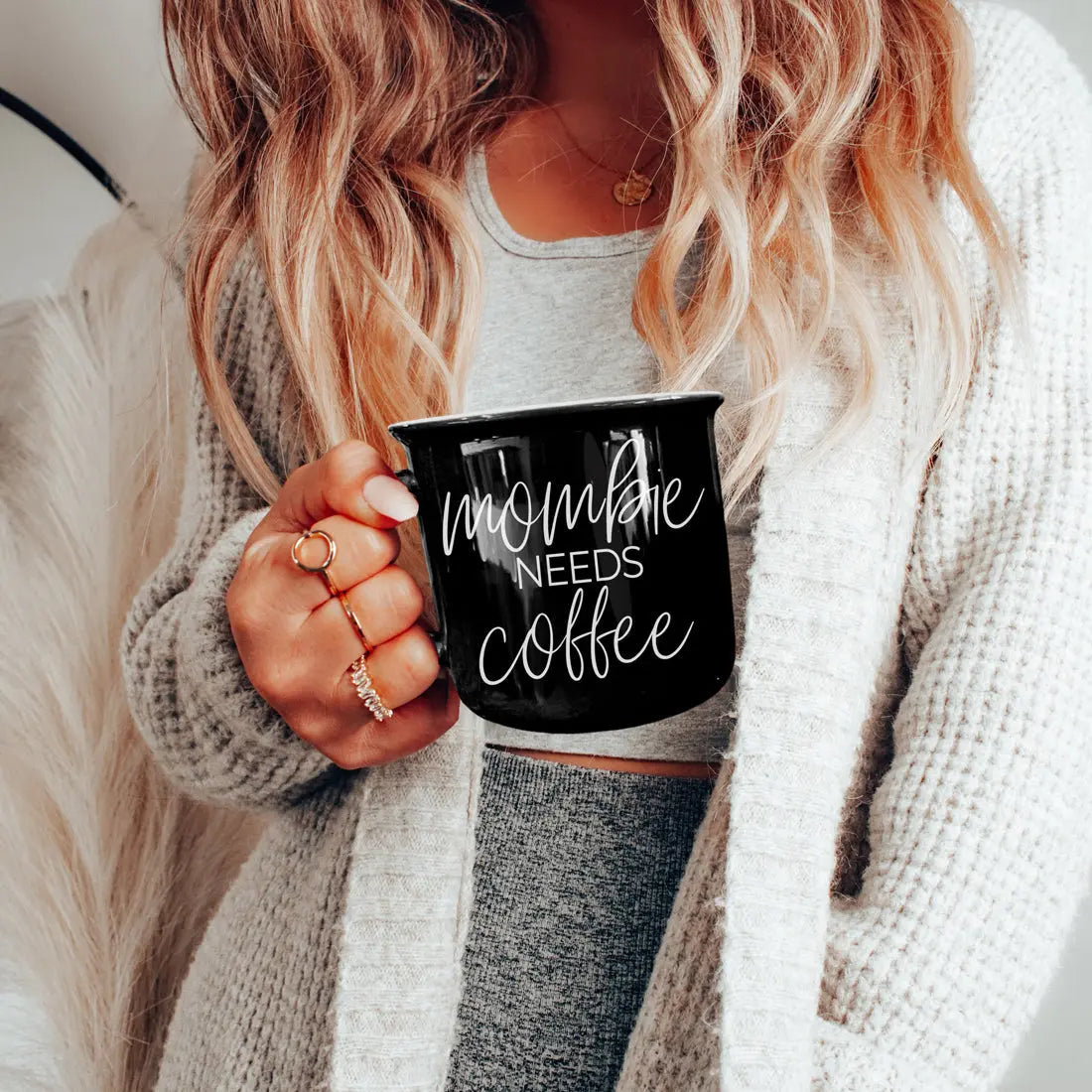 Person holding a black mug with 'Creepy + Spooky Before Coffee' text, wearing a light gray sweater and dark pants.