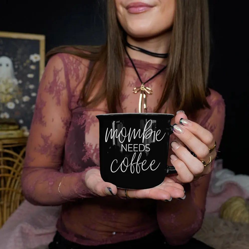 Person holding a black mug with 'mommy needs coffee' text in a cozy setting.