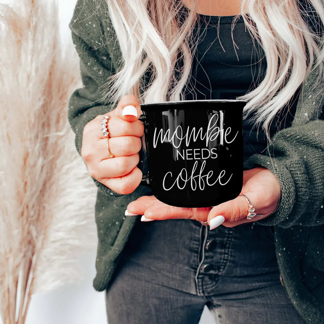 Person holding a black mug with 'mom life needs coffee' text, wearing a green jacket and black top.
