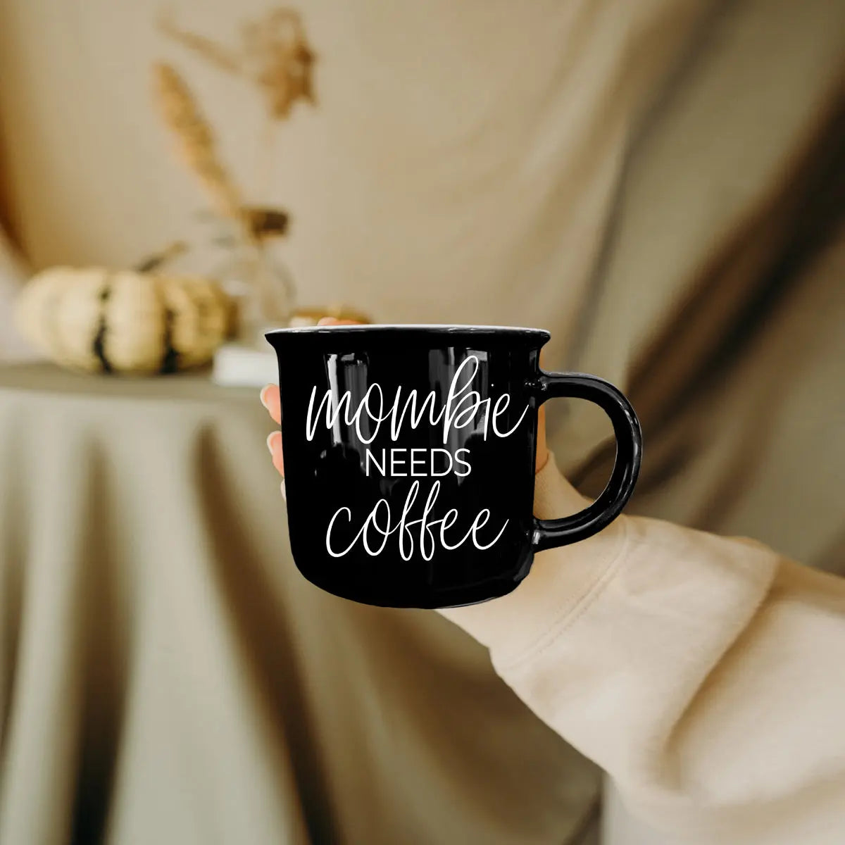 Black mug with 'mommy needs coffee' text held by a person in a cozy indoor setting.