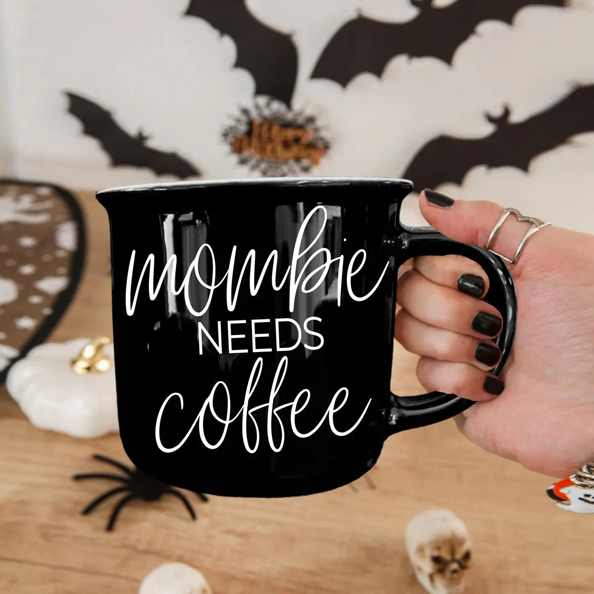 Black mug with 'mombie needs coffee' text held by a hand, Halloween decorations in the background