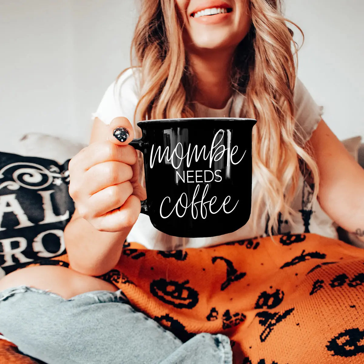 Person holding a black mug with 'mommy needs coffee' text, wearing an orange Halloween-themed blanket.
