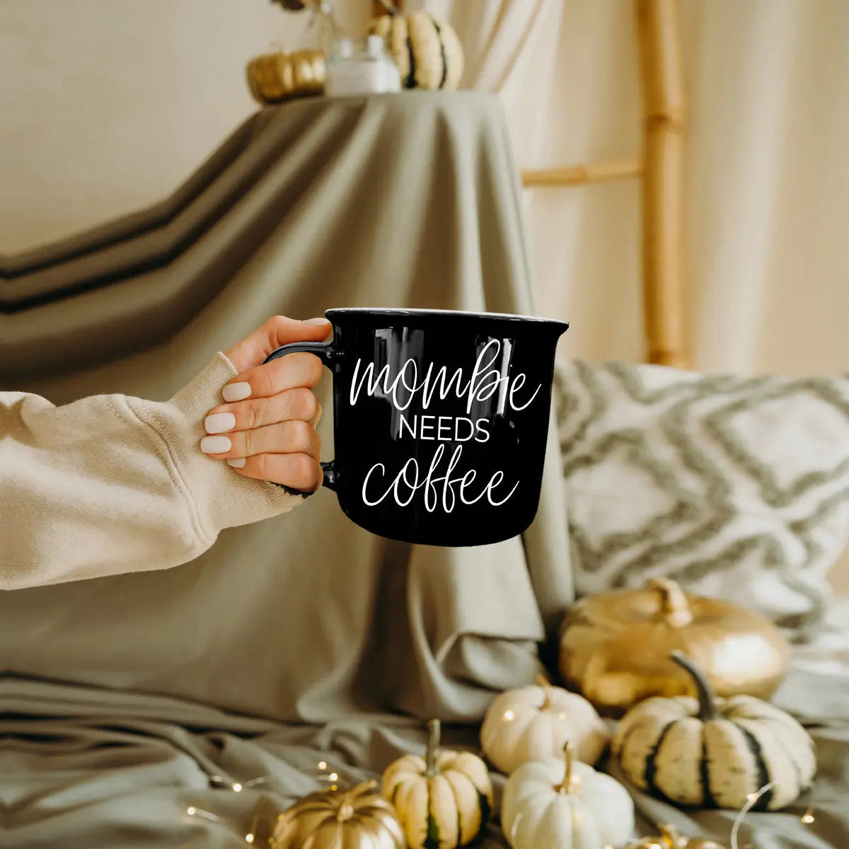 Person holding a black mug with 'Mommy Needs Coffee' text, surrounded by pumpkins and cozy decor.