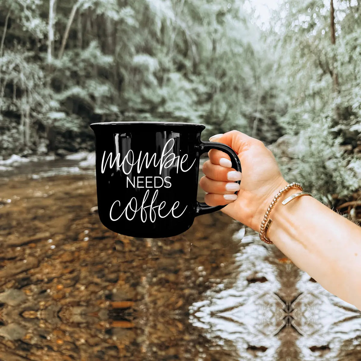Hand holding a black mug with 'mommy needs coffee' text against a forest background