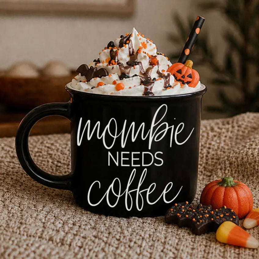 Black mug with 'mombie needs coffee' text, topped with Halloween-themed whipped cream and candy, on a textured surface.