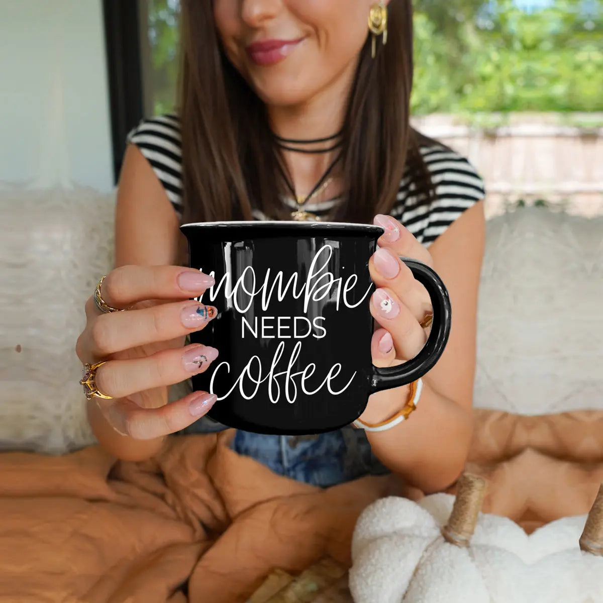 Woman holding a black mug with 'Mommy Needs Coffee' text, sitting on a couch outdoors.