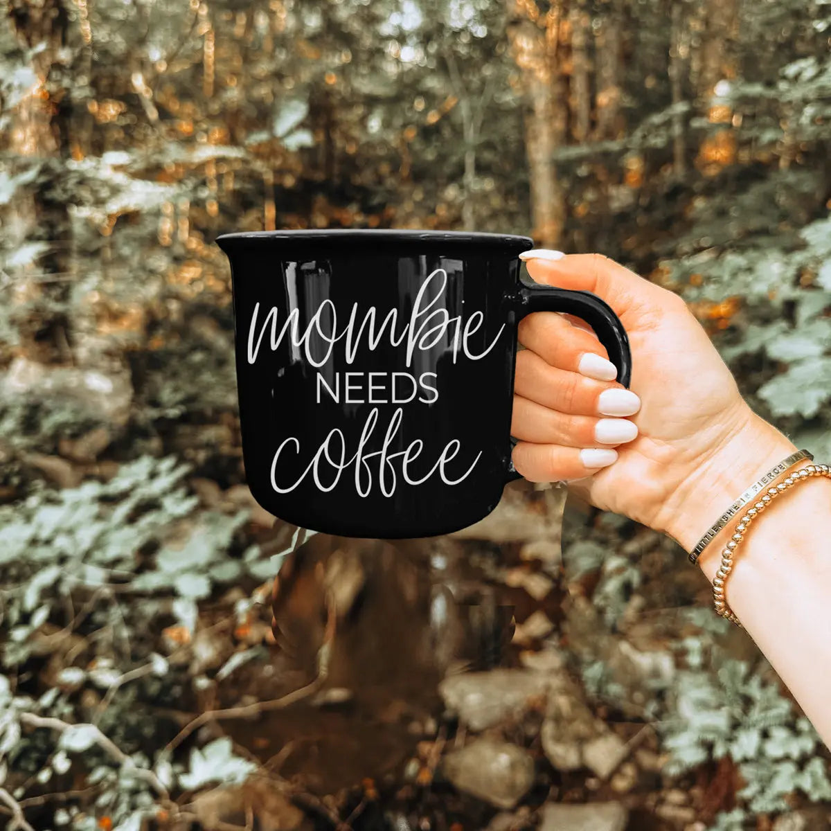 Hand holding a black mug with 'mommy needs coffee' text against a forest background