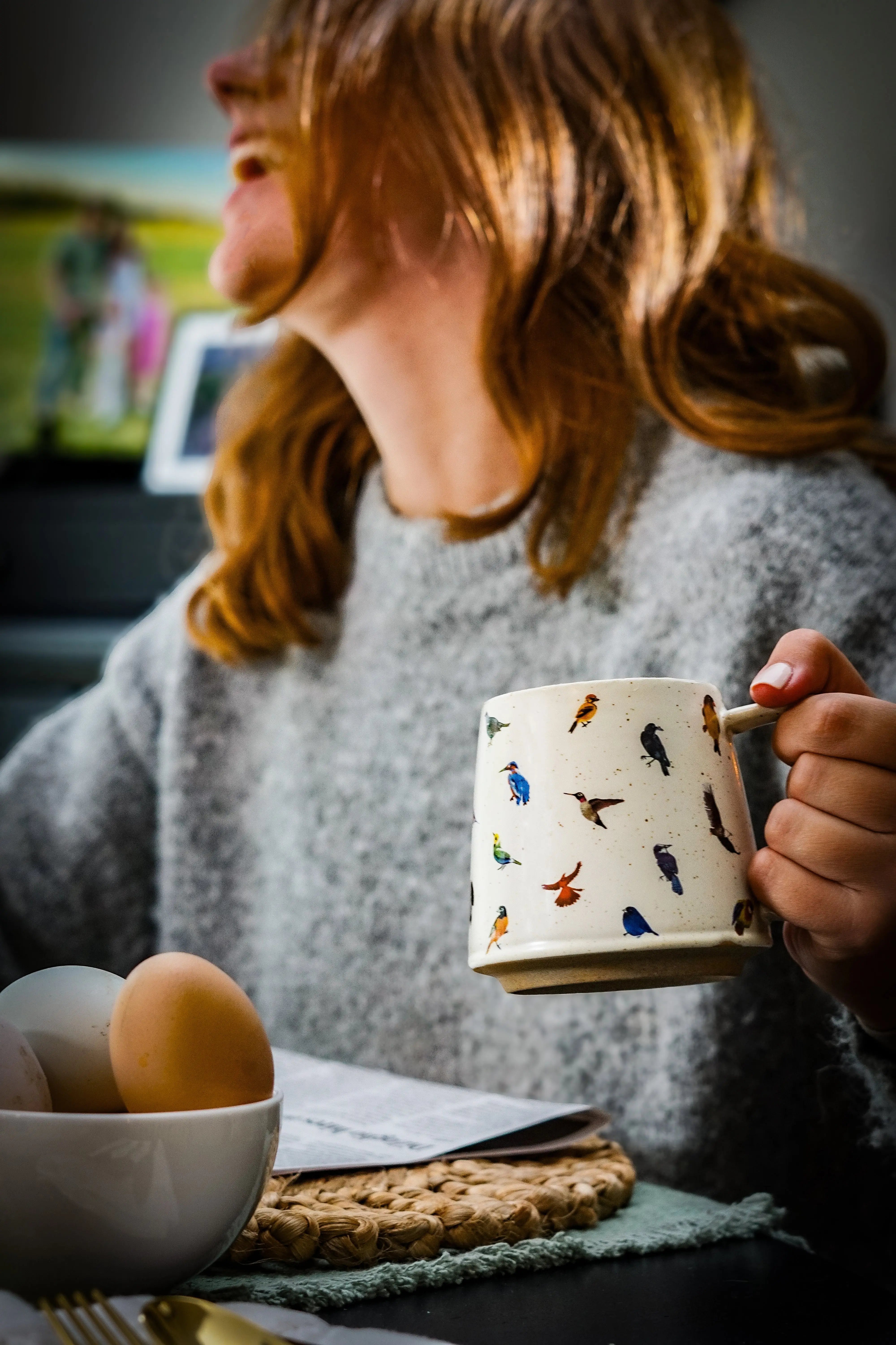 Woman holding bird-patterned mug, cozy farmhouse kitchen scene with eggs and woven placemat