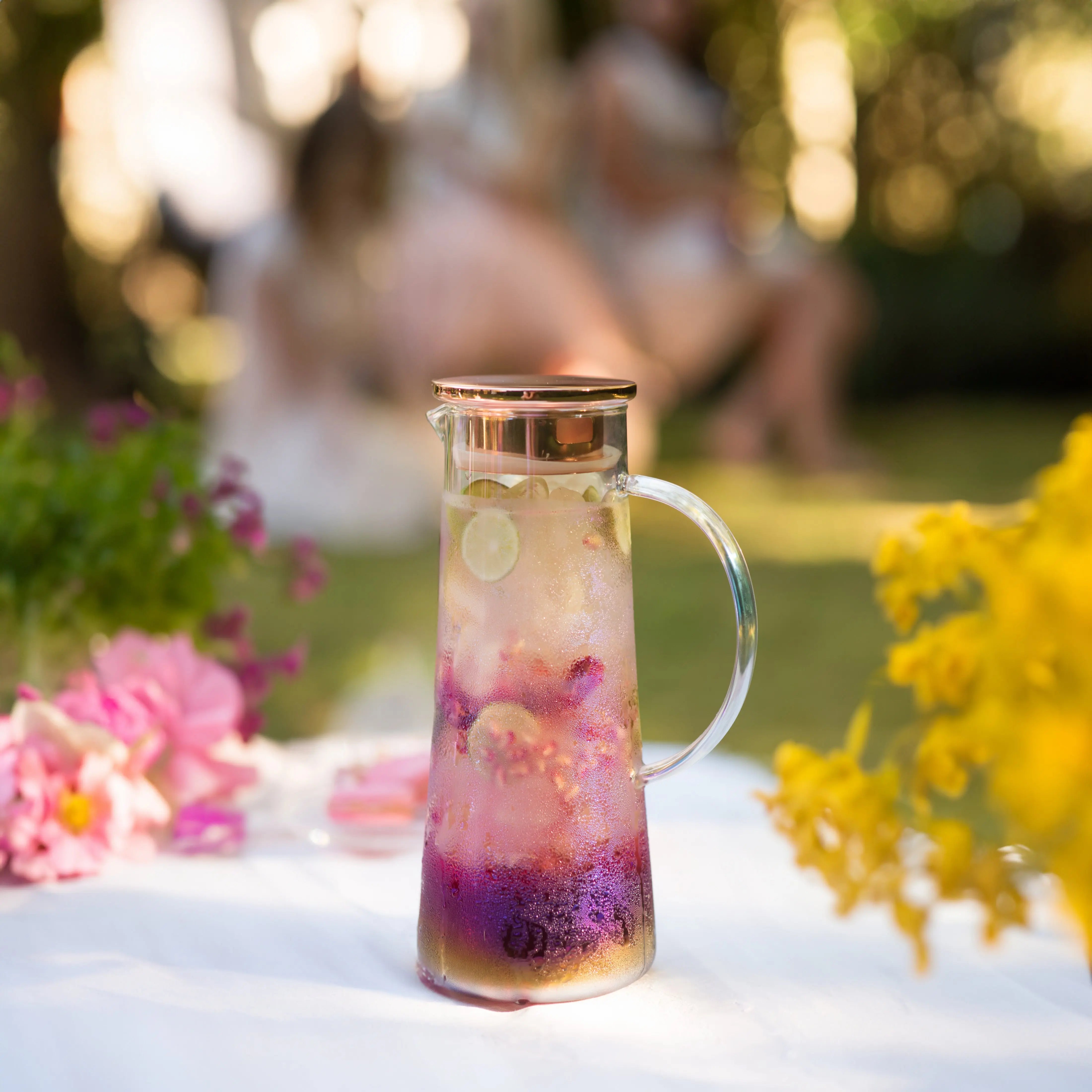 Glass carafe with floral infusion on a table outdoors, surrounded by flowers.