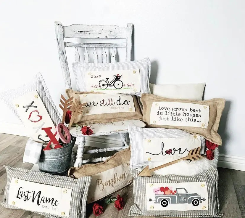 A styled collection of rustic decorative throw pillows arranged on a white chair and floor against a white wall. The pillows feature various designs including embroidered text ('we still do', 'love grows best in little houses just like this.