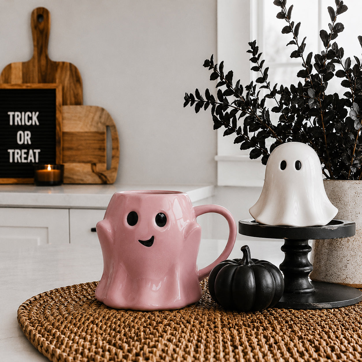 Pink ghost-shaped mug on a table with Halloween decorations including a black pumpkin and a sign saying 'Trick or Treat'.