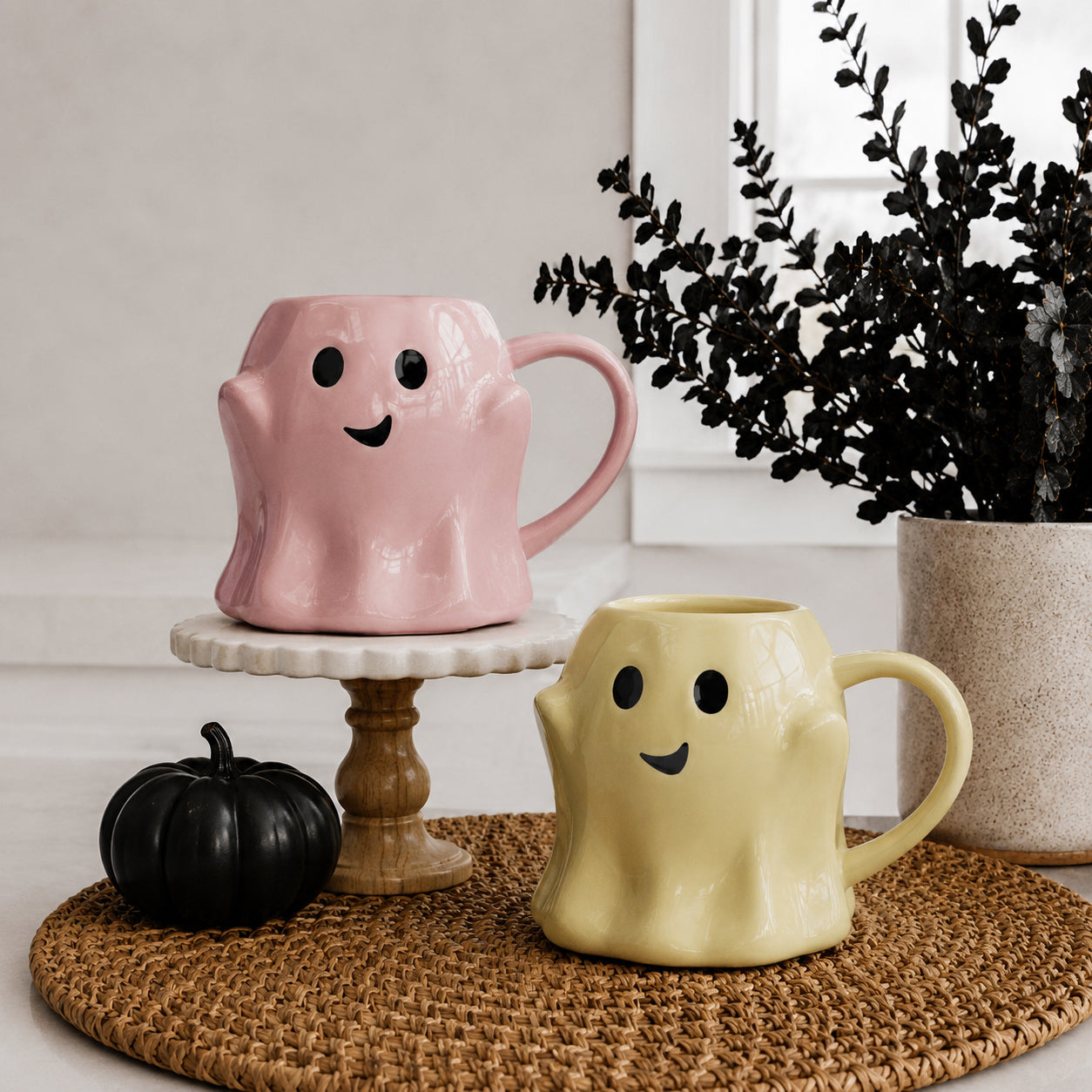 Two ghost-shaped mugs, one pink and one yellow, on a woven mat with a black pumpkin and plant in the background.
