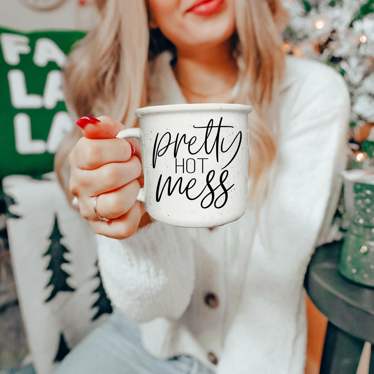 Person holding a mug with 'pretty hot mess' text in a cozy indoor setting.