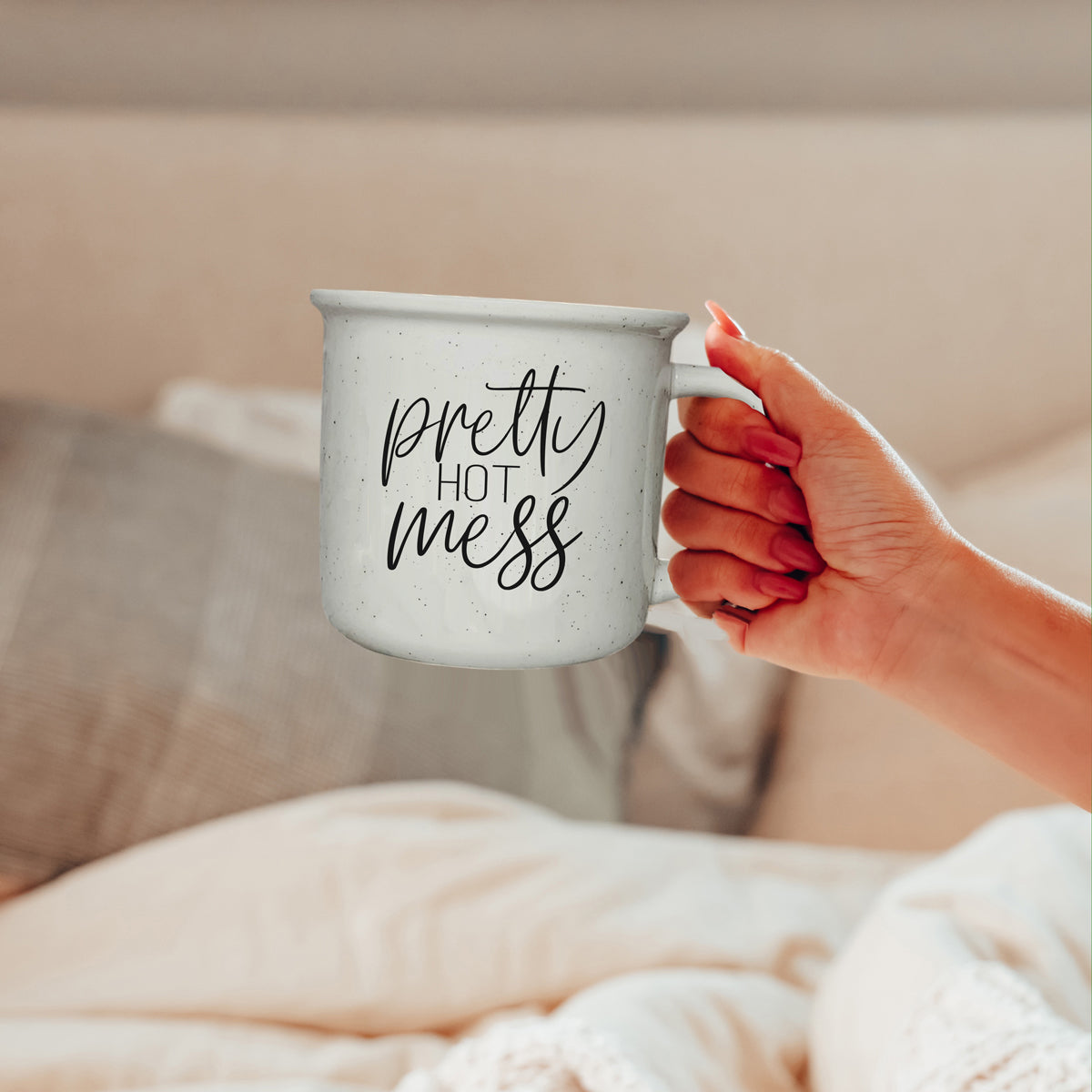 Hand holding a mug with 'pretty hot mess' text in a cozy indoor setting.