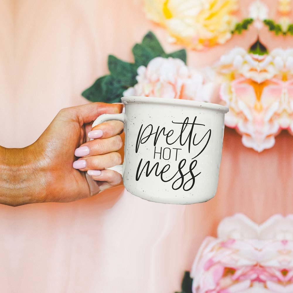Hand holding a mug with 'pretty hot mess' text against a floral background