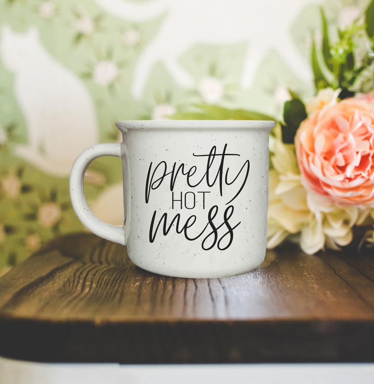 White mug with 'Pretty Hot Mess' text on a wooden surface with flowers in the background
