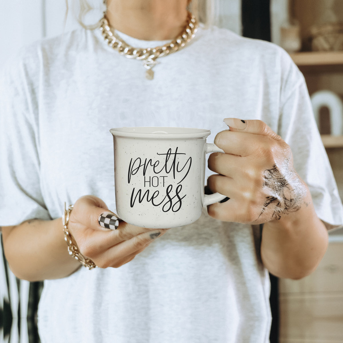Person holding a mug with 'pretty hot mess' text, wearing a white shirt and gold jewelry.