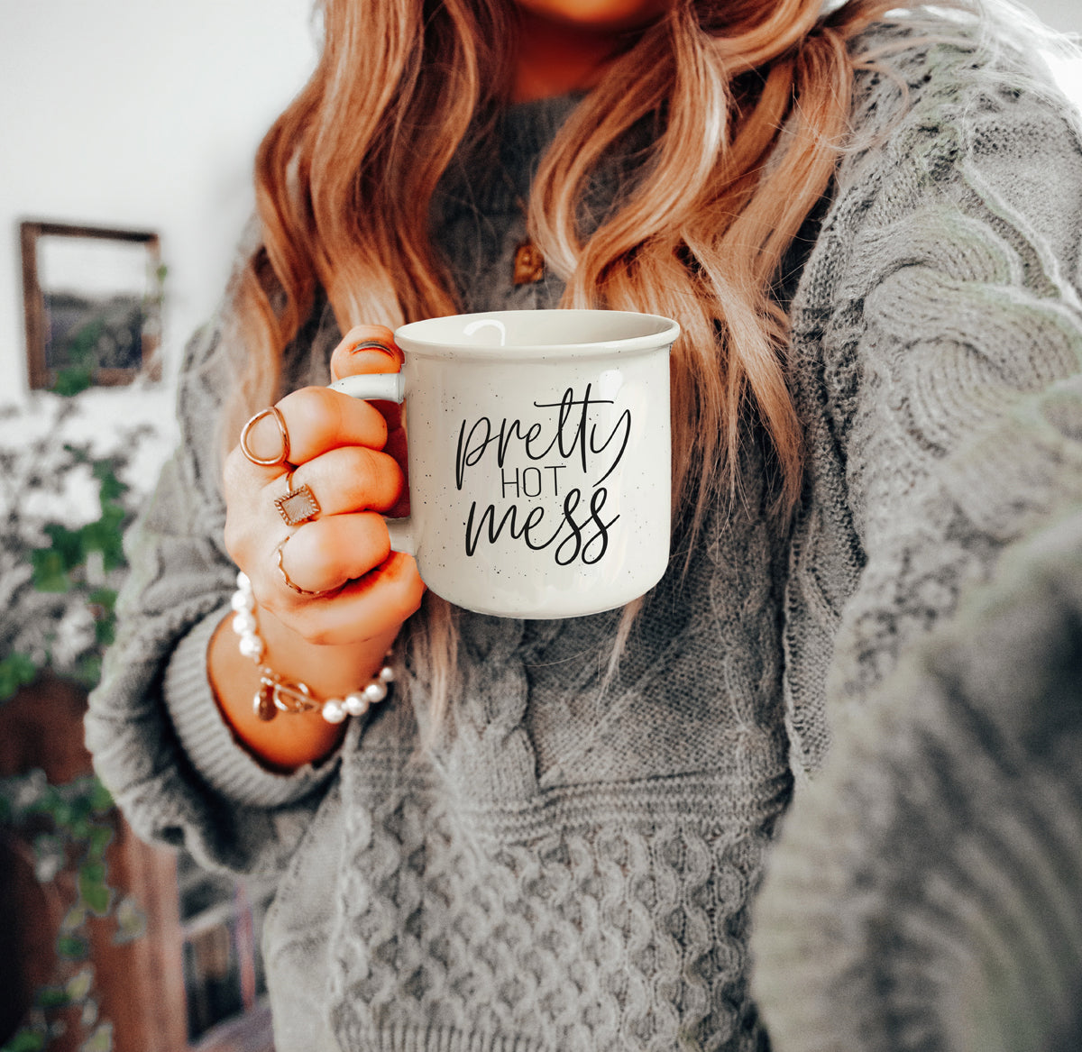 Person holding a mug with text, wearing a gray sweater indoors.