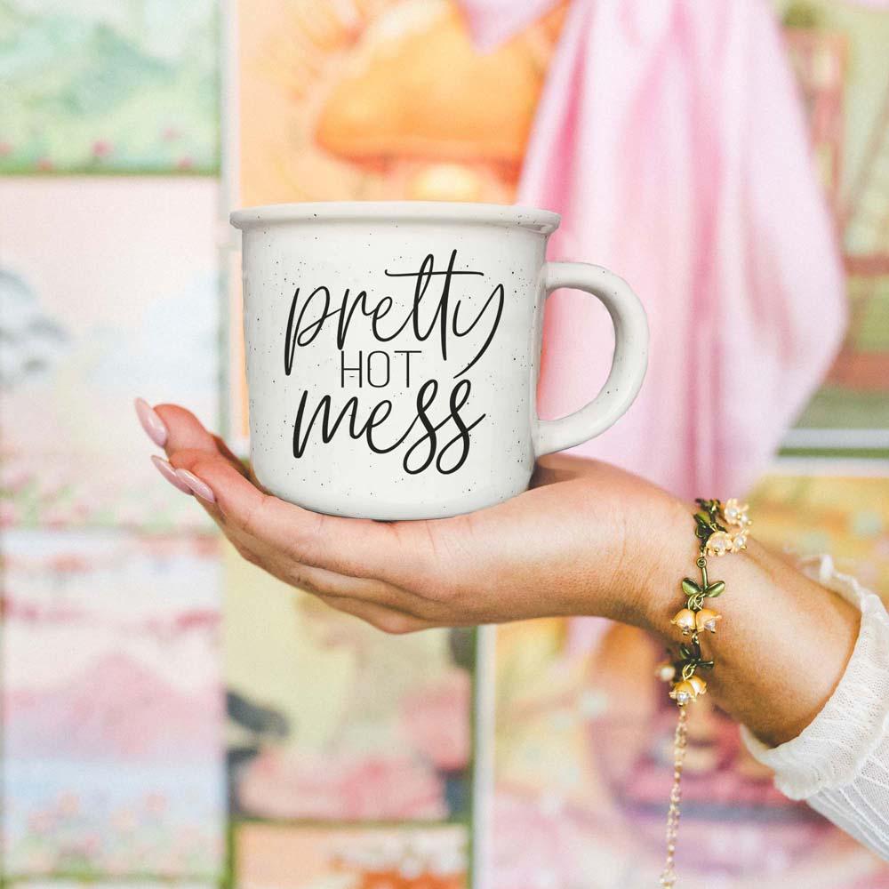 Hand holding a mug with 'pretty hot mess' text against a blurred background