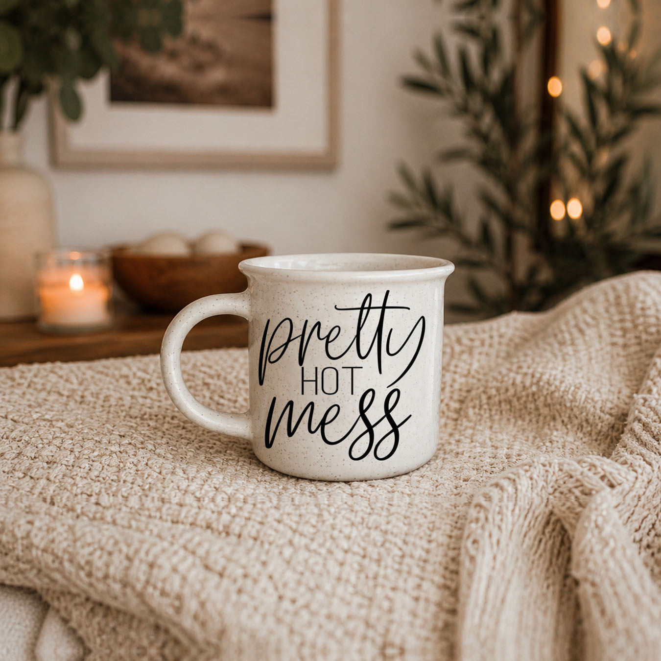 White mug with 'pretty hot mess' text on a textured surface with candles and plants in the background