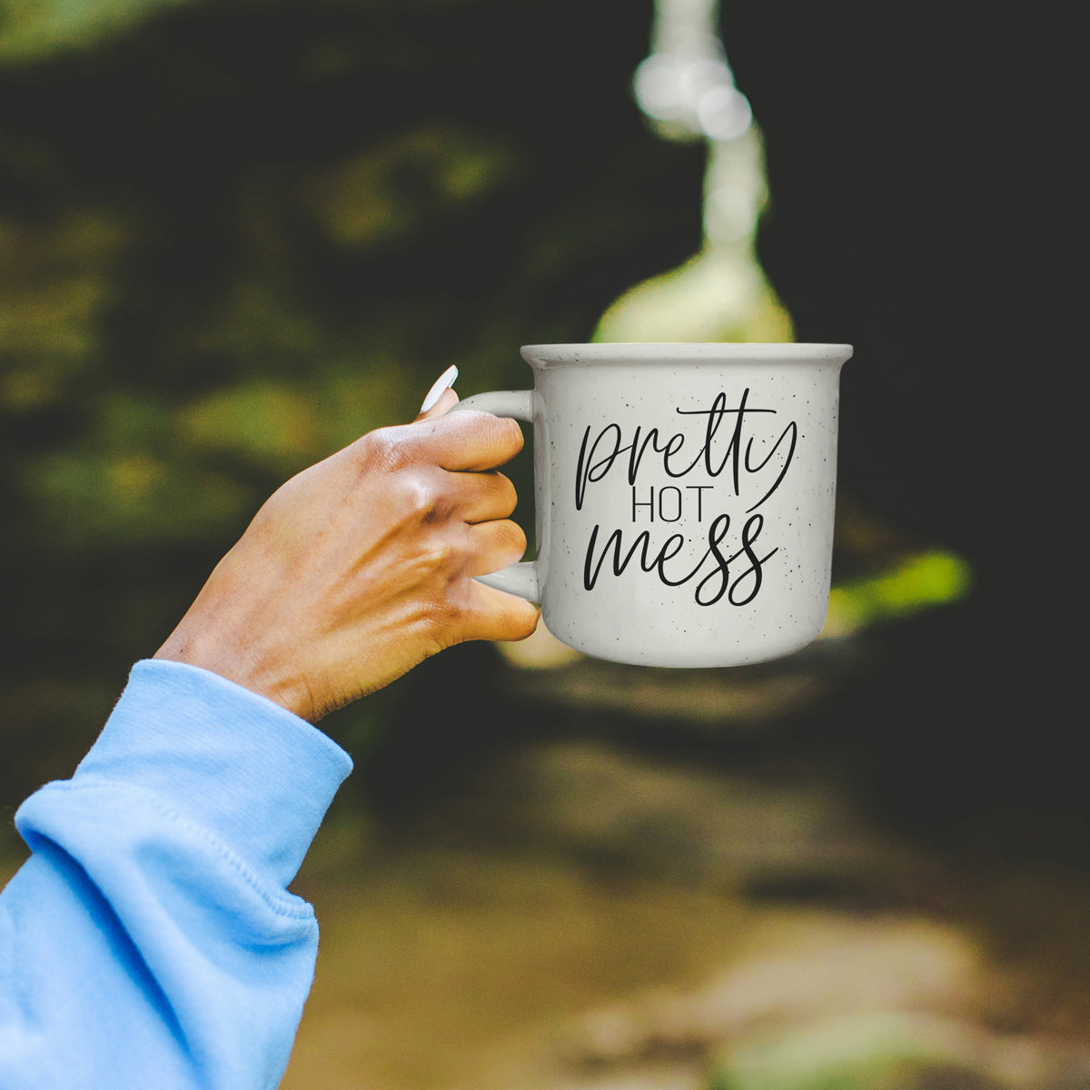 Hand holding a mug with 'Pretty Hot Mess' text against a blurred natural background