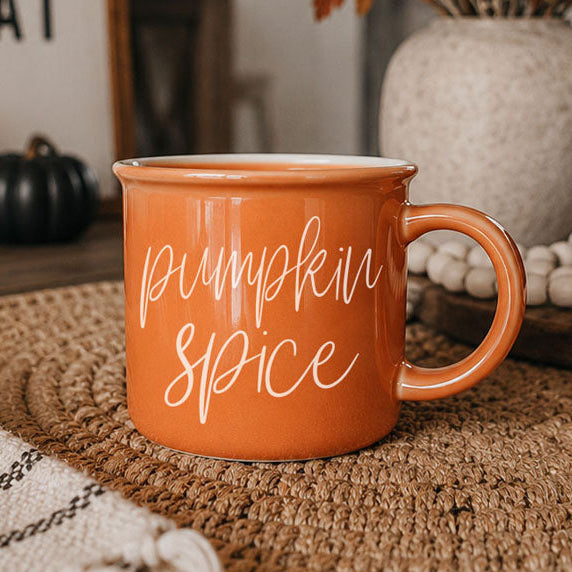 An orange ceramic mug with 'pumpkin spice' written in white script, sitting on a woven placemat in a cozy, autumn-themed setting.