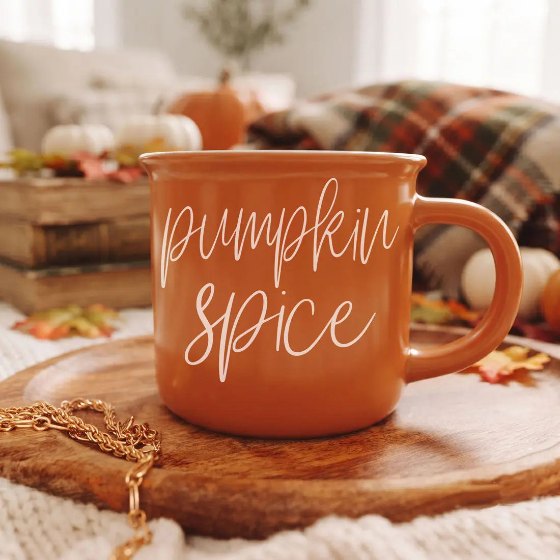 An orange ceramic mug with 'pumpkin spice' written in white script, resting on a wooden board in a cozy autumn-themed setting with pumpkins, a plaid blanket, and a gold chain.