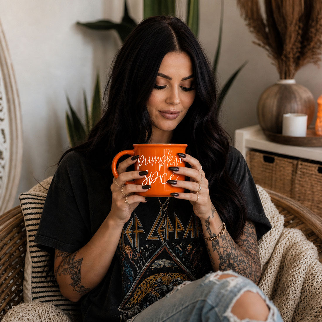 A woman with long black hair sits in a woven chair holding an orange ceramic mug with 'pumpkin spice' written on it in white script. She is wearing a black graphic t-shirt and ripped light-wash jeans in a cozy indoor setting with plants and home decor in the background.