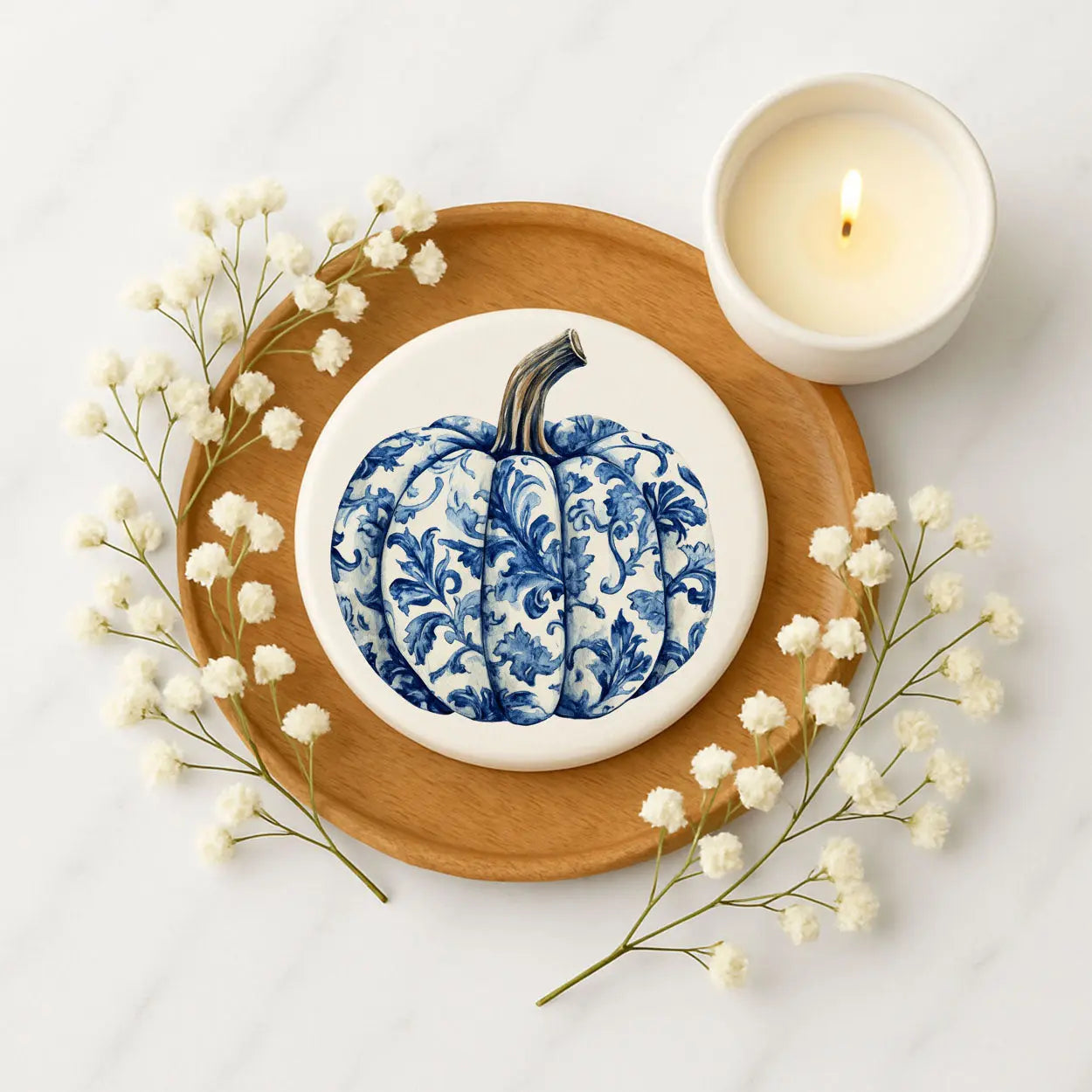 Decorative blue and white pumpkin on a white plate with a wooden tray, candles, and flowers on a light background