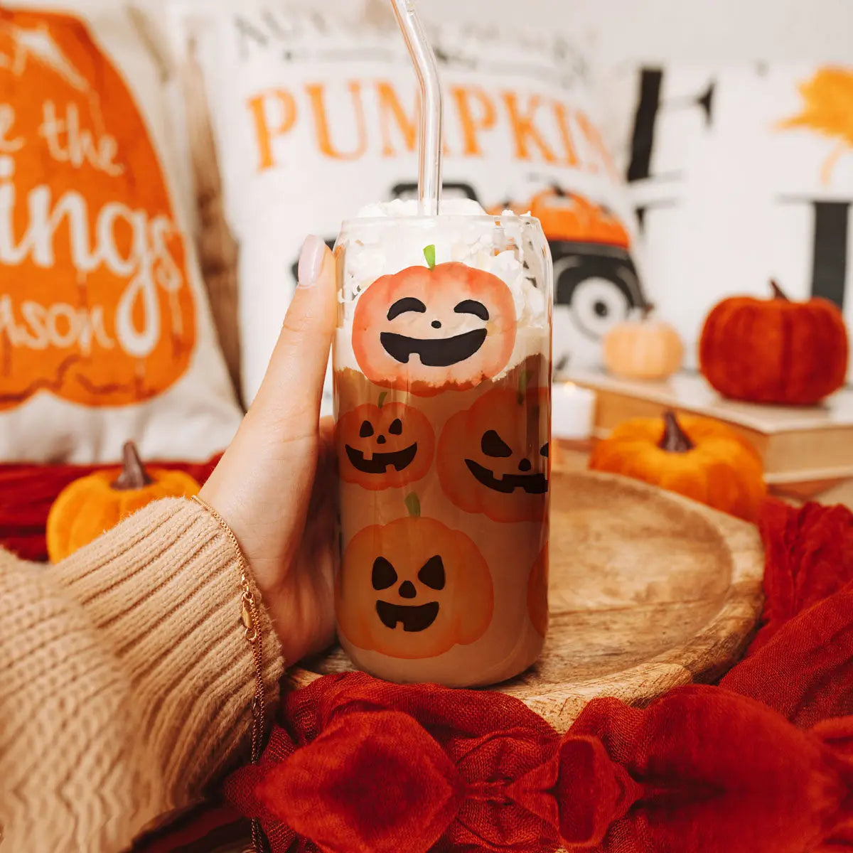 Hand holding a Halloween-themed cup with pumpkins and a straw, surrounded by autumn decorations.