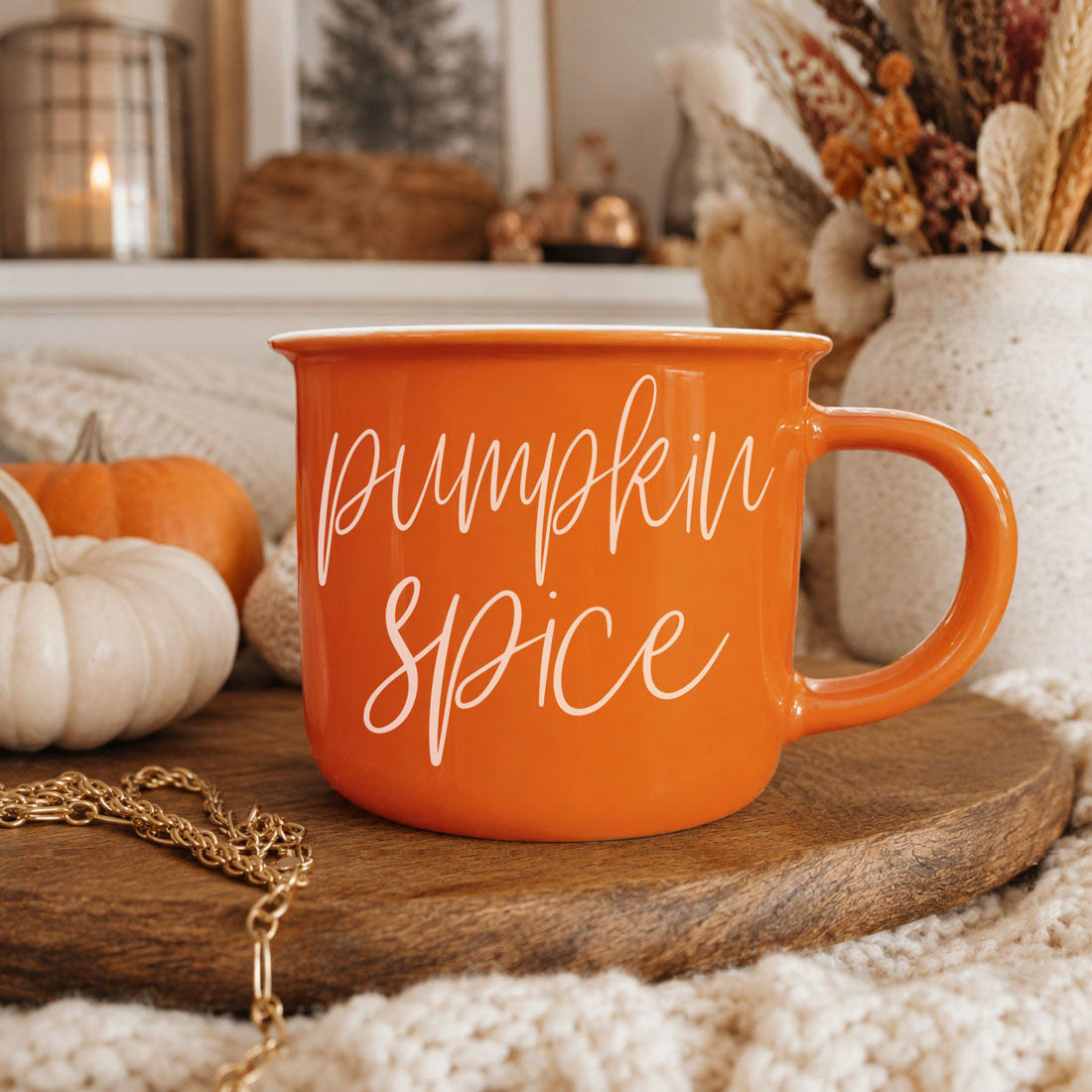 An orange ceramic mug with 'pumpkin spice' written in white script, displayed on a wooden tray in a cozy autumn-themed setting with pumpkins, a knit blanket, and dried flowers.