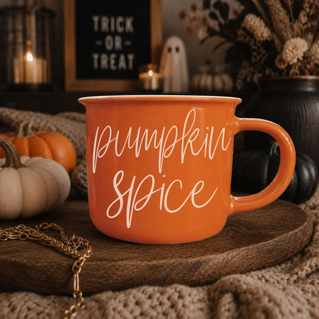 An orange ceramic mug with 'pumpkin spice' written in white script, displayed on a wooden tray in a cozy autumn setting with pumpkins, candles, and a sign reading 'Trick or Treat'.