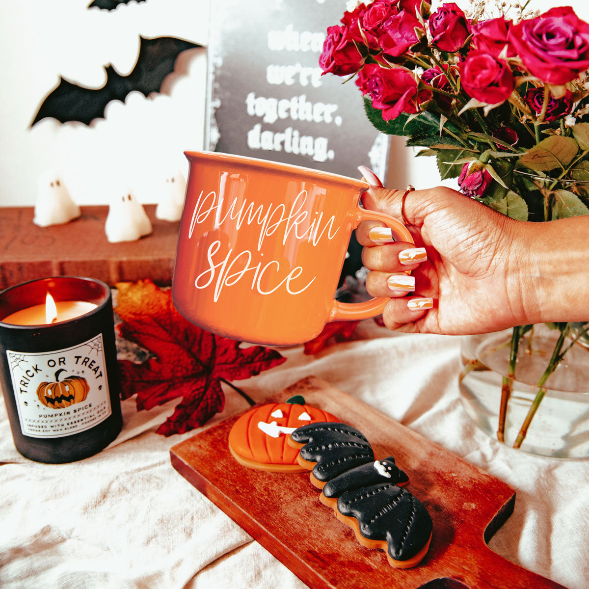 Person holding an orange mug with Halloween-themed text, surrounded by festive items like a candle and cookies.